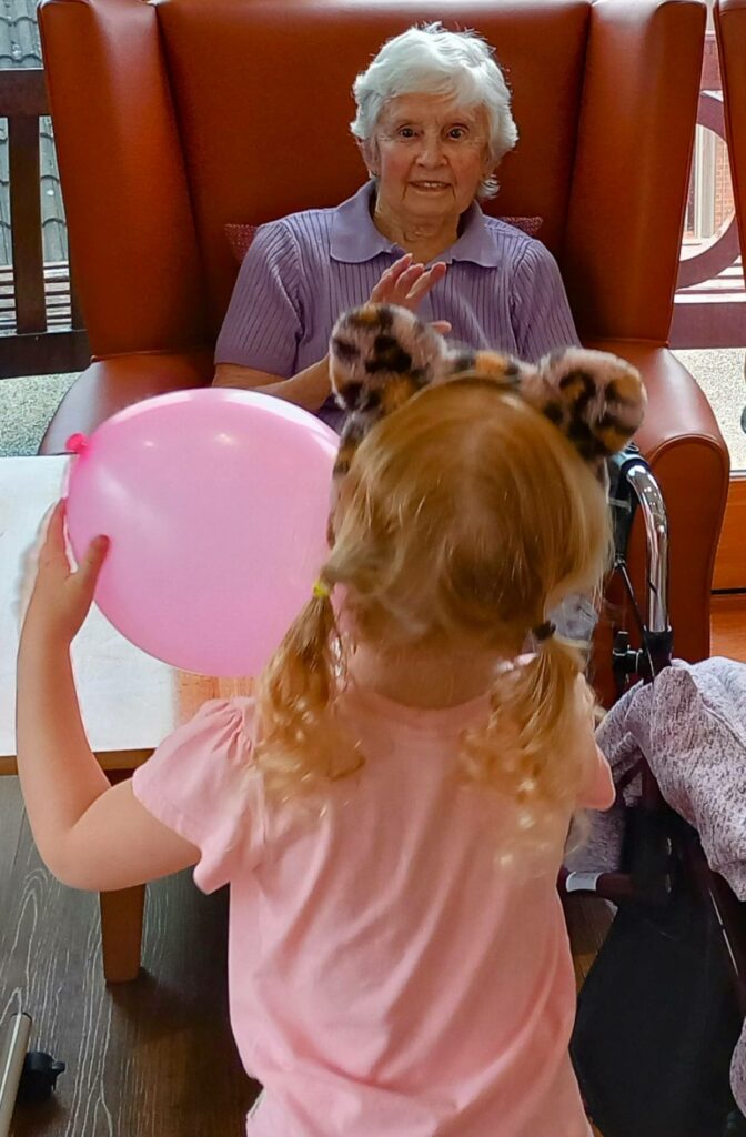 An elderly woman sits in an armchair at a residential care home, smiling and waving at a young girl holding a pink balloon and wearing animal ear headbands, sharing a moment of intergenerational fun. Heres the revised version in British English: An elderly woman sits in an armchair at a residential care home, smiling and waving to a young girl holding a pink balloon and wearing animal ear headbands, sharing an intergenerational moment of fun.