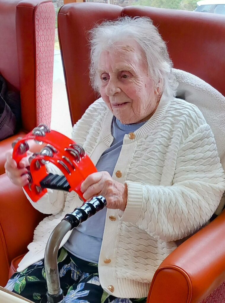 An elderly woman in a residential care home sits in an armchair, holding a red tambourine and smiling. She is wearing a white jumper and has a walking frame in front of her.