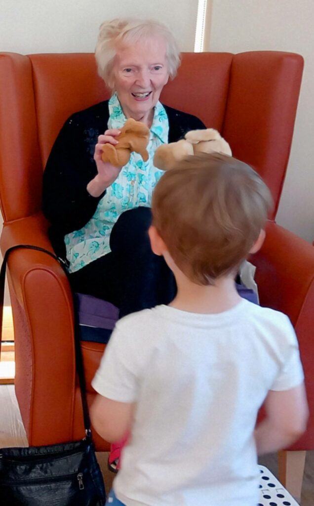 An elderly woman in a residential care home sits in an armchair, holding up two small soft toys and smiling at a young child, sharing a moment of intergenerational fun.