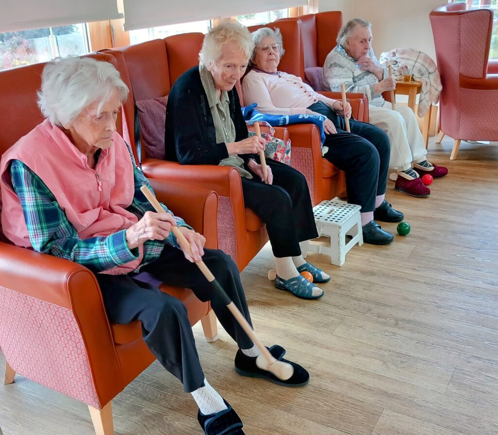 Four elderly women enjoy an active afternoon in armchairs—Carla massages her foot with a wooden tool whilst the others relax with blankets. Mobility aids and therapy balls encourage getting active together.