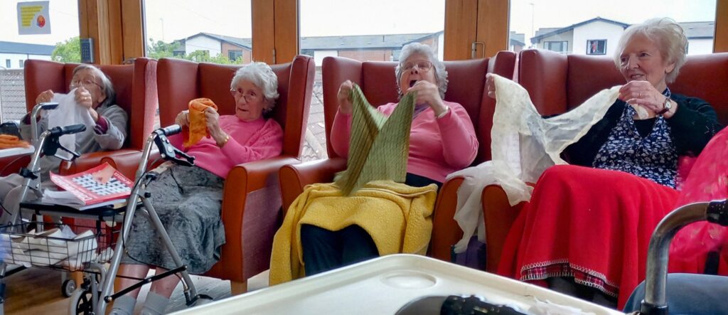 Four elderly women sit in high-backed chairs, each holding or folding pieces of knitting or fabric, in a bright communal room at a residential care home.