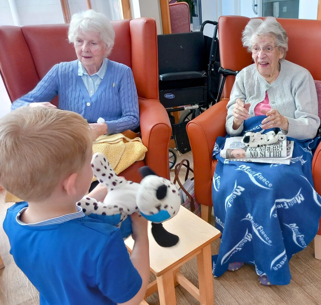 A young boy shares intergenerational fun by showing a stuffed Dalmatian toy to two elderly women sitting in armchairs at a care home; one holds a similar soft toy and a newspaper.