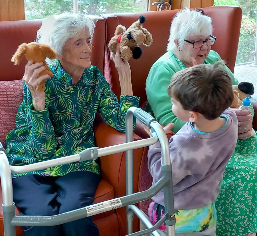 Two elderly women sit in armchairs holding soft toy dogs, enjoying intergenerational fun with a young boy standing in front of them. A walking frame is visible in the foreground.