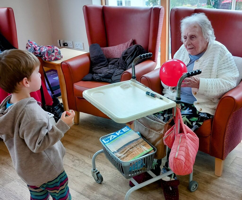 An elderly woman sits in an armchair with a red balloon and a walking frame tray as a young child stands in front of her, enjoying intergenerational activities in the well-lit residential care home filled with armchairs.