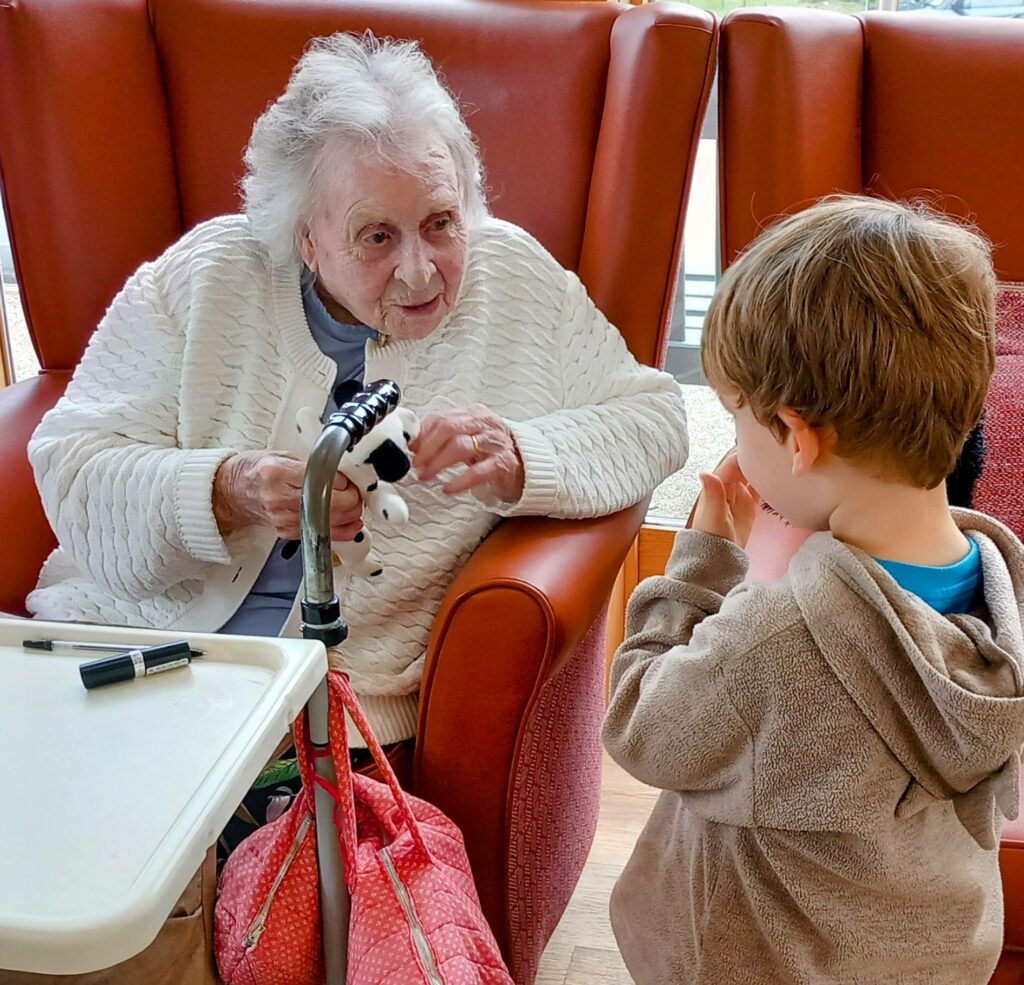 An elderly woman in a care home sits in an armchair, holding a small toy as she enjoys intergenerational fun with a young boy standing nearby. A table with personal belongings is beside her.