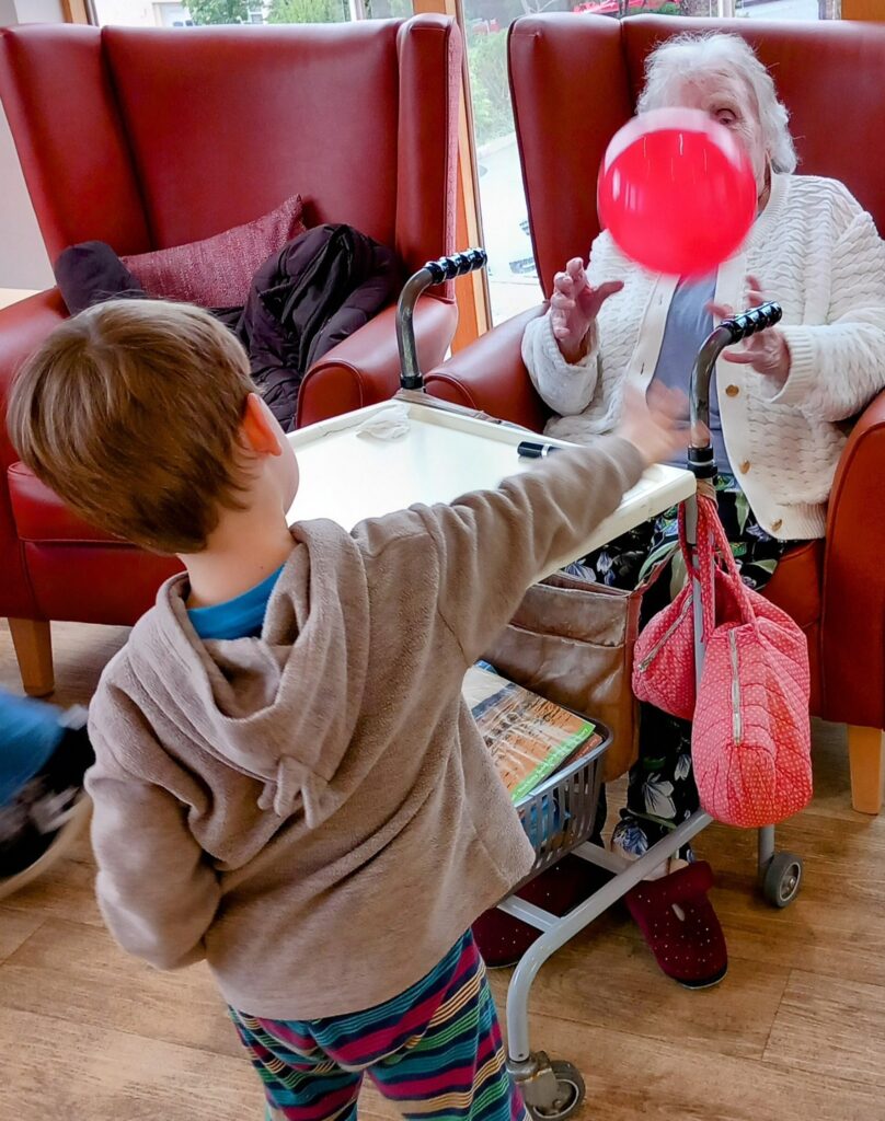 A young boy plays with a red balloon, reaching towards an elderly woman seated in a chair with a walking frame in front of her, capturing heartwarming intergenerational fun in a residential care home.