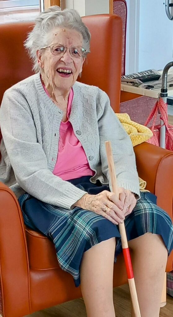 Carla, an elderly woman with glasses and grey hair, sits smiling in an orange armchair, holding a wooden stick—ready for an active afternoon in her cardigan, pink shirt and checked skirt.