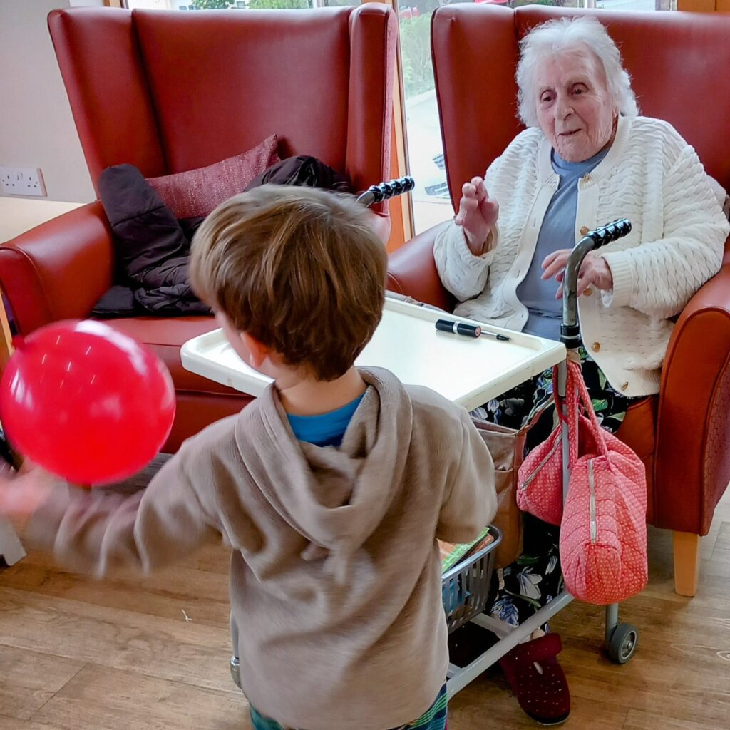A young boy enjoys intergenerational fun with a red balloon in front of an elderly woman seated with her walking frame and tray table at a residential care home.