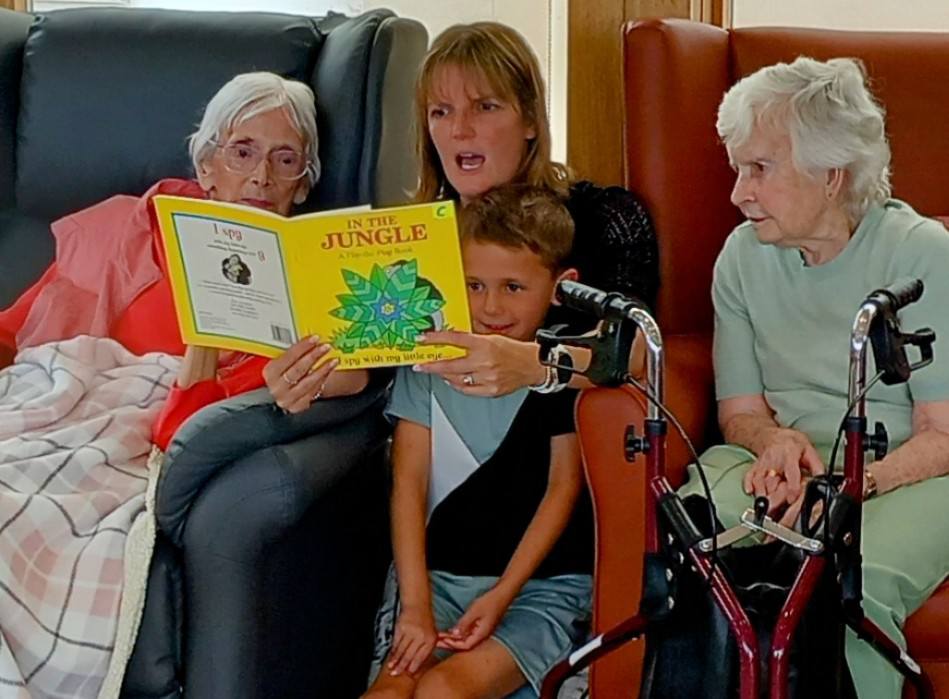 A woman reads a book titled In the Jungle to a young boy, with two elderly women sitting beside them, one using a walking frame.