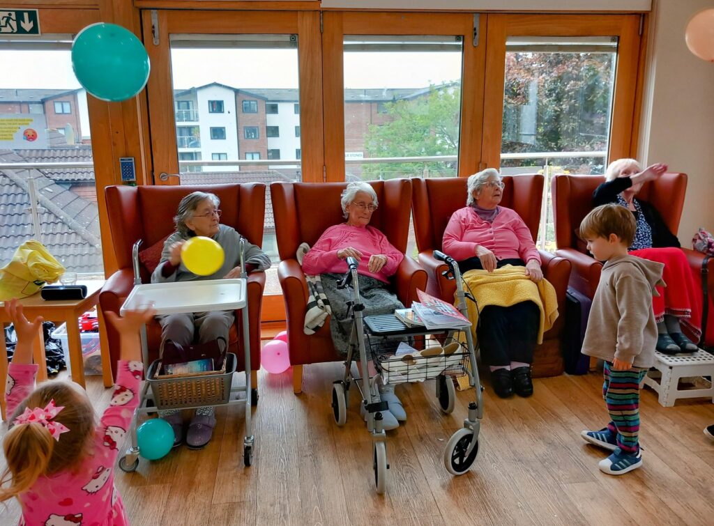 Four elderly women sit in armchairs by large windows in a residential care home, while children with balloons enjoy intergenerational fun nearby. A walking frame and small table are in front of the women.