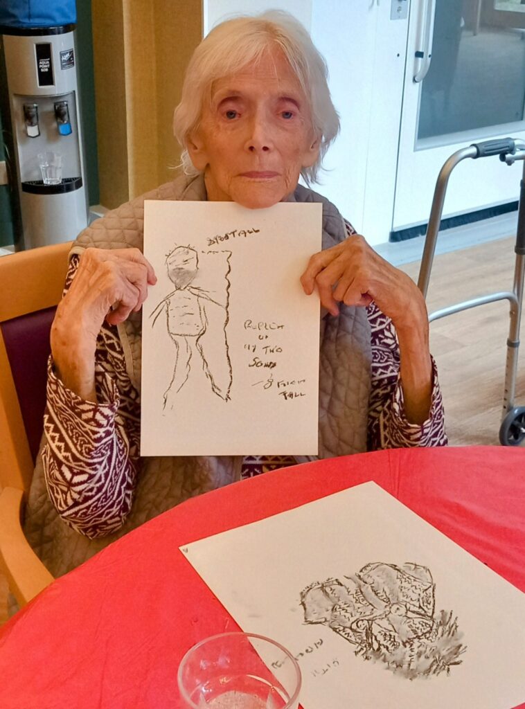 An elderly woman sits at a table, holding up a pencil drawing, with another sketch on the table in front of her. A walking frame and water cooler are visible—just another joyful June Crafternoon filled with Creative Sparks.