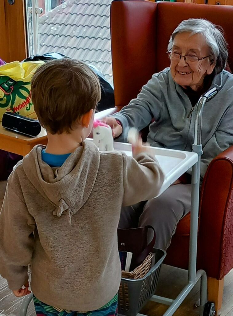 An elderly woman in a residential care home smiles warmly as a young boy stands before her, holding a toy—capturing a moment of intergenerational fun.