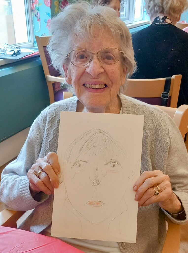 An elderly woman with glasses smiles while holding a pencil-drawn portrait, seated at a table in a brightly lit room during Crafternoon, where Creative Sparks ignite her artistic expression.