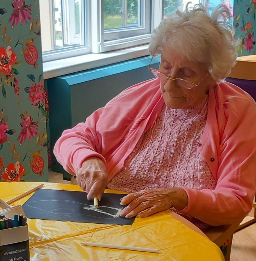 An elderly woman enjoys a Crafternoon in June, drawing with white chalk on black paper in a brightly lit, floral-decorated room, letting her creative sparks shine.