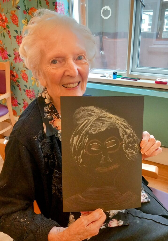 An elderly woman sits indoors, smiling and holding up a black sheet of paper with a white chalk drawing of a face—her creative spark shining during a joyful Crafternoon June event.