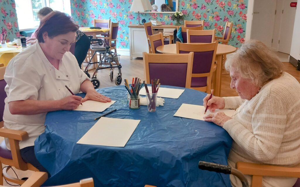 Two women sit at a table covered with a blue cloth, sharing giggles and creative sparks as they draw with coloured pencils in a brightly decorated room during a June Crafternoon.