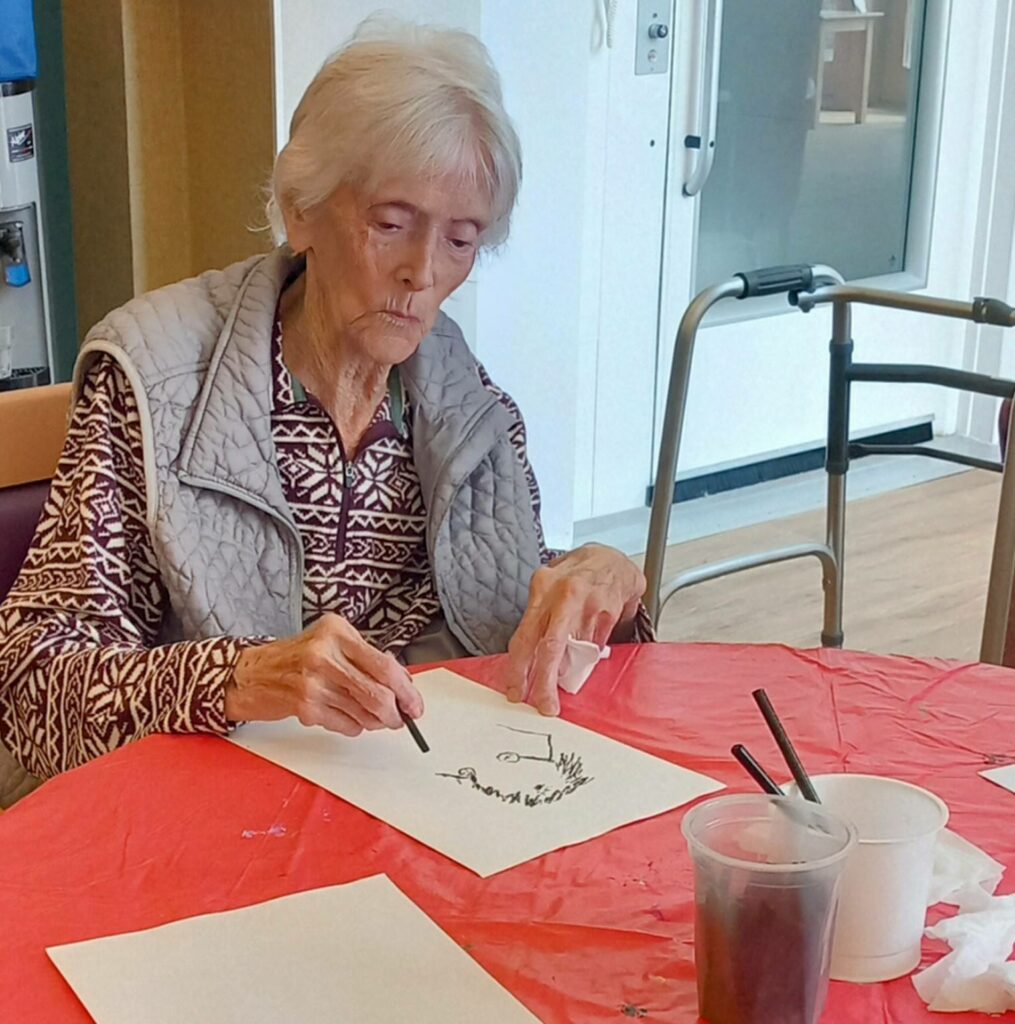 An elderly woman sits at a table during June’s Crafternoon, concentrating on painting a design onto white paper. Art supplies and cups are on the red tablecloth, and a walking frame is visible in the background.