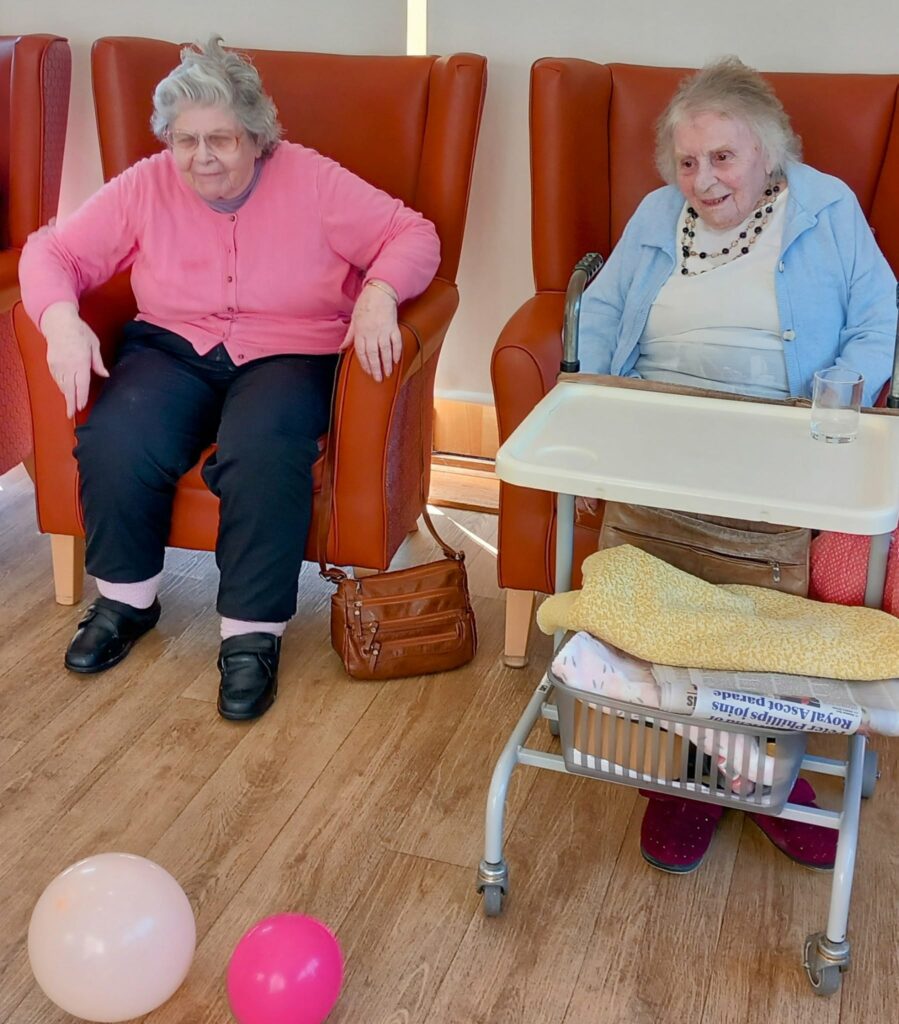 Two elderly women sit in armchairs at a care home; one uses a walking frame with a tray holding a glass of water, blanket, and newspaper. Balloons on the floor suggest recent intergenerational fun.