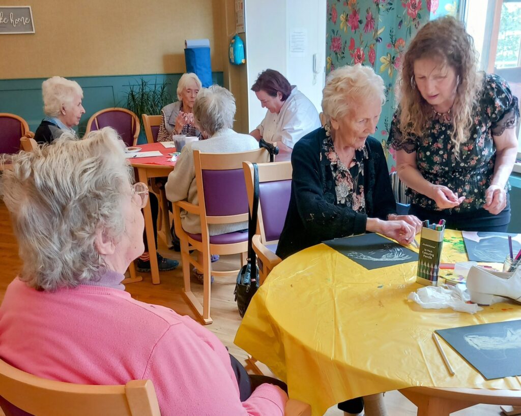 A group of elderly women sit around tables in a brightly lit room, taking part in arts and crafts activities with an instructor during a lively June Crafternoon.