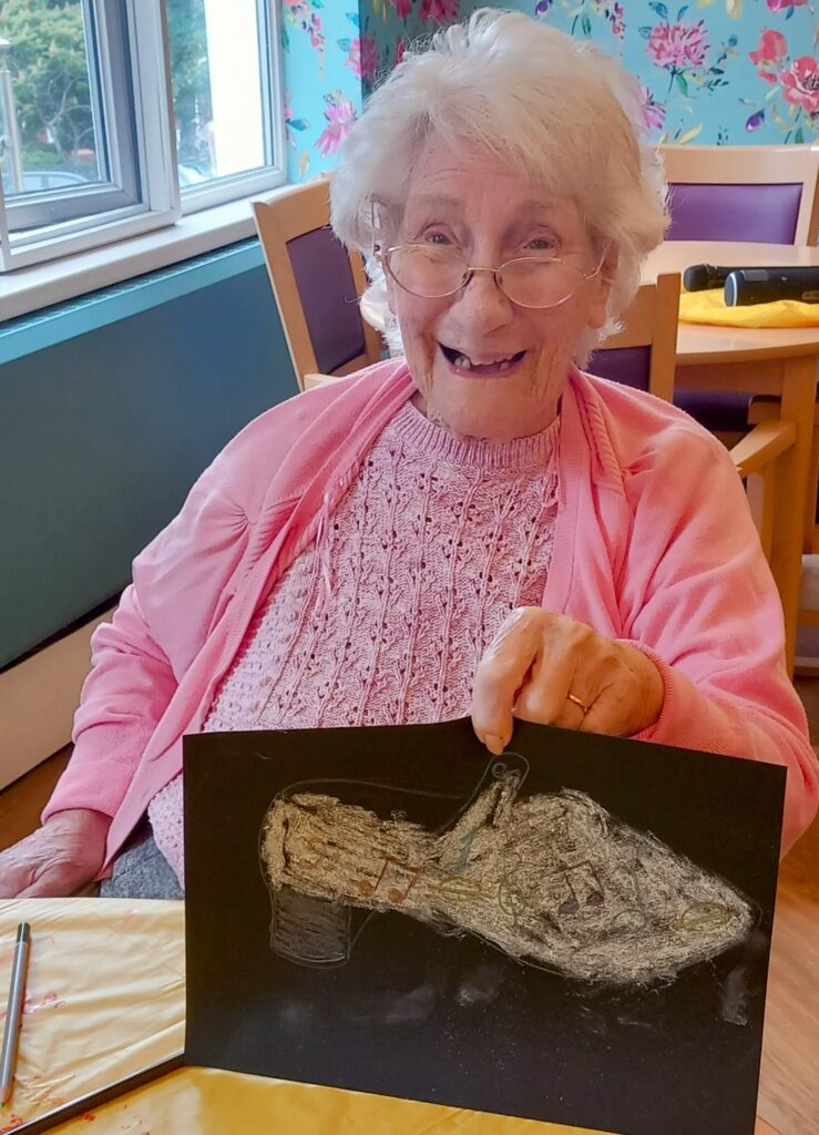 An elderly woman wearing a pink jumper and glasses smiles while holding up a drawing of a shoe during the June Crafternoon, surrounded by Creative Sparks in a brightly lit room.