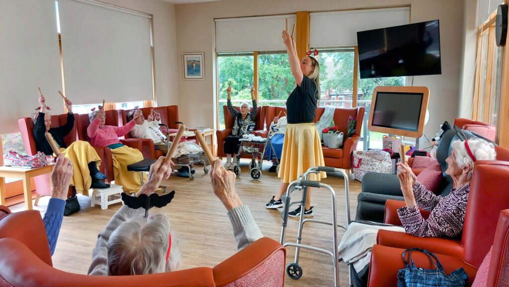 A group of elderly people in a care home lounge share laughter as they follow an instructor in a yellow skirt, raising one arm together during a lively activity.