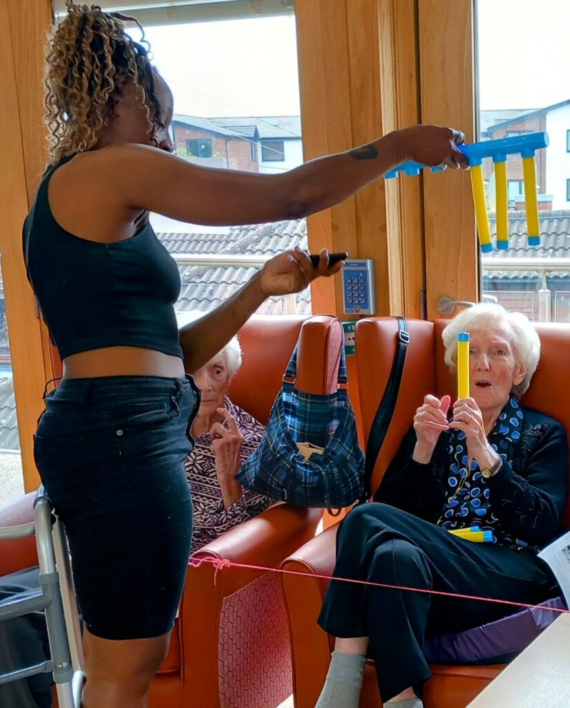A woman dressed in black assists two elderly women seated in armchairs with a colourful activity involving yellow and blue plastic tubes and string.