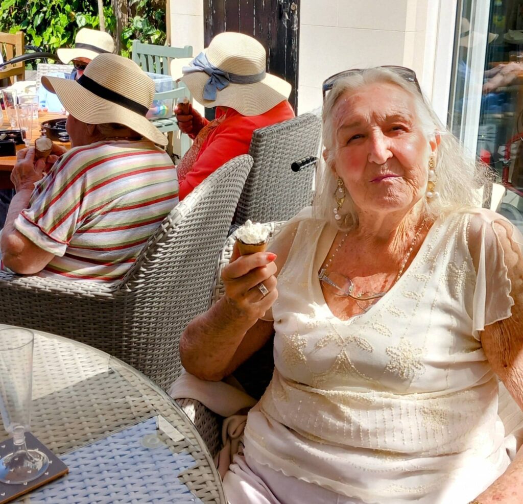 An elderly woman sits at an outdoor table holding an ice cream cone during Vera’s Special Day, while other women in sunhats enjoy the garden party in the background.