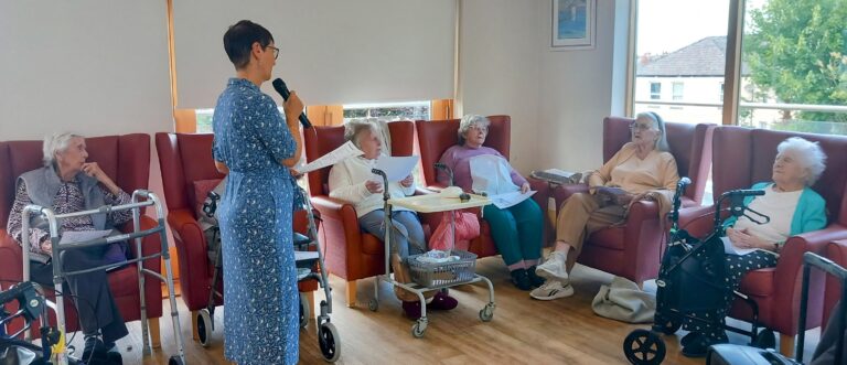 A woman with a microphone stands before five elderly women seated in armchairs, holding papers as sunlight streams in—walking frames nearby hint at gentle strolls or trips down memory lane.