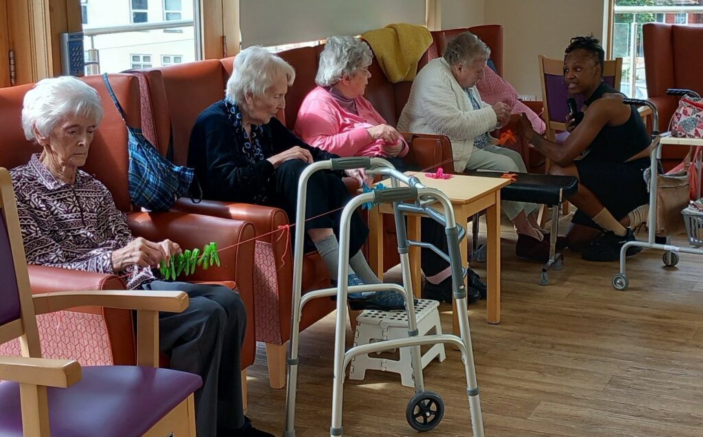 Four elderly women sit in armchairs engaging in craft activities, while a staff member chats with one of them in a bright, communal room. Walking frames are nearby.