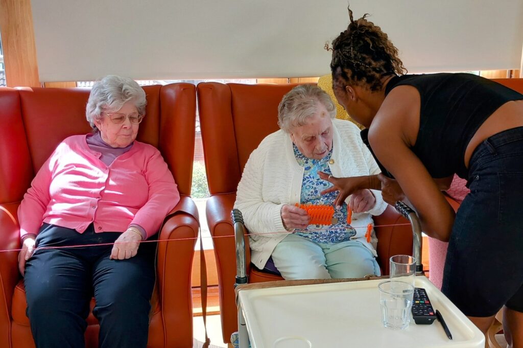 Two elderly women are seated in armchairs; one is knitting while a woman dressed in black leans in to offer assistance or engage her in conversation. A tray with a glass and remote control sits in the foreground.