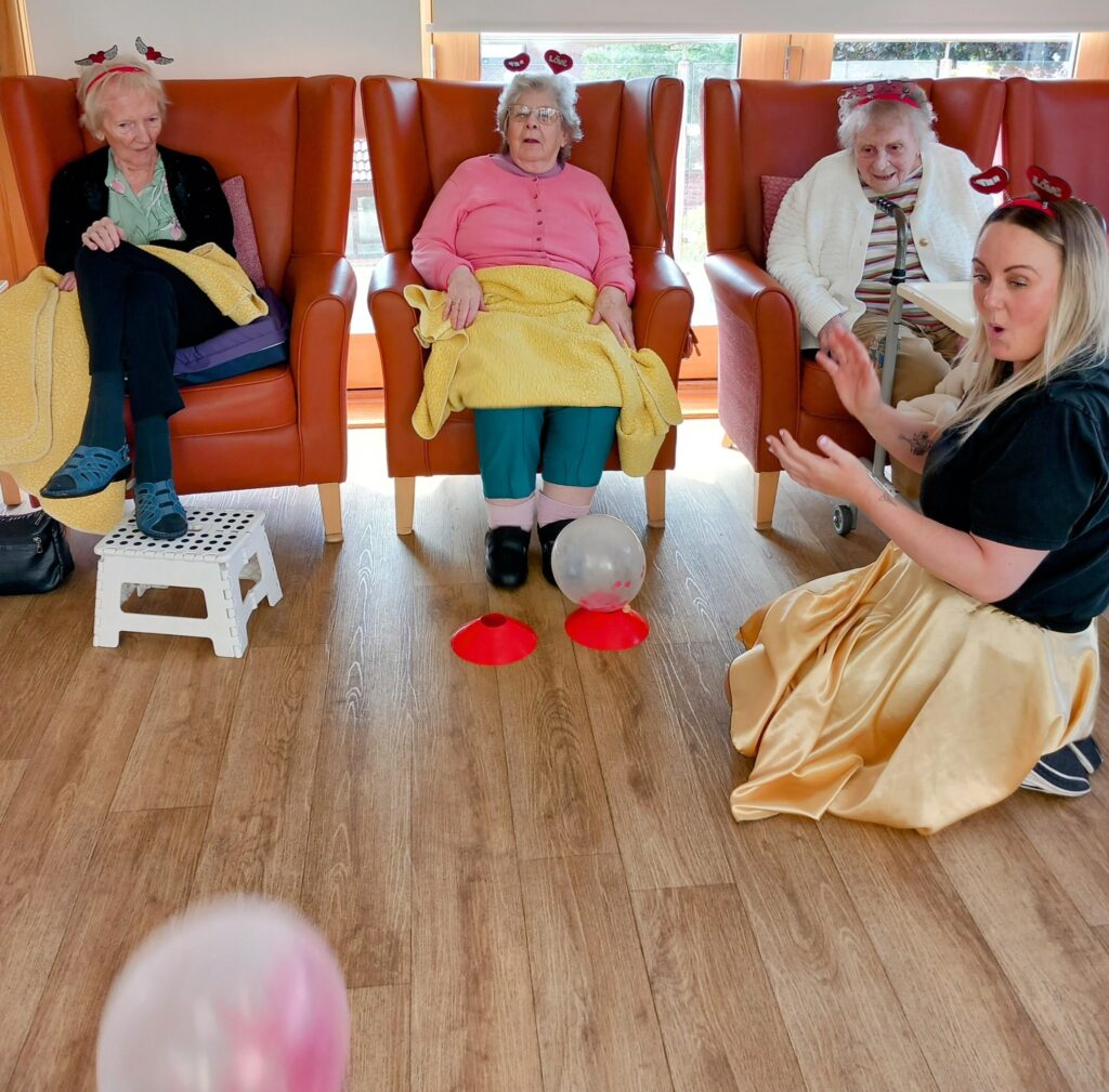 Three elderly women sit in chairs wearing heart headbands, watching Jenna kneeling on the floor as she plays with balloons in a brightly lit room filled with laughter and love. (In this sentence, there is little to change between American and British English as the vocabulary used is common to both variants. The primary differences between American and British English often involve words like colour/colour or theater/theatre, which are not present here. If you’d like more distinctly British phrasing, you could consider something like:) Three elderly ladies sit in chairs, each wearing a heart-shaped headband, as they watch Jenna kneel on the floor playing with balloons in a brightly lit room full of laughter and affection.