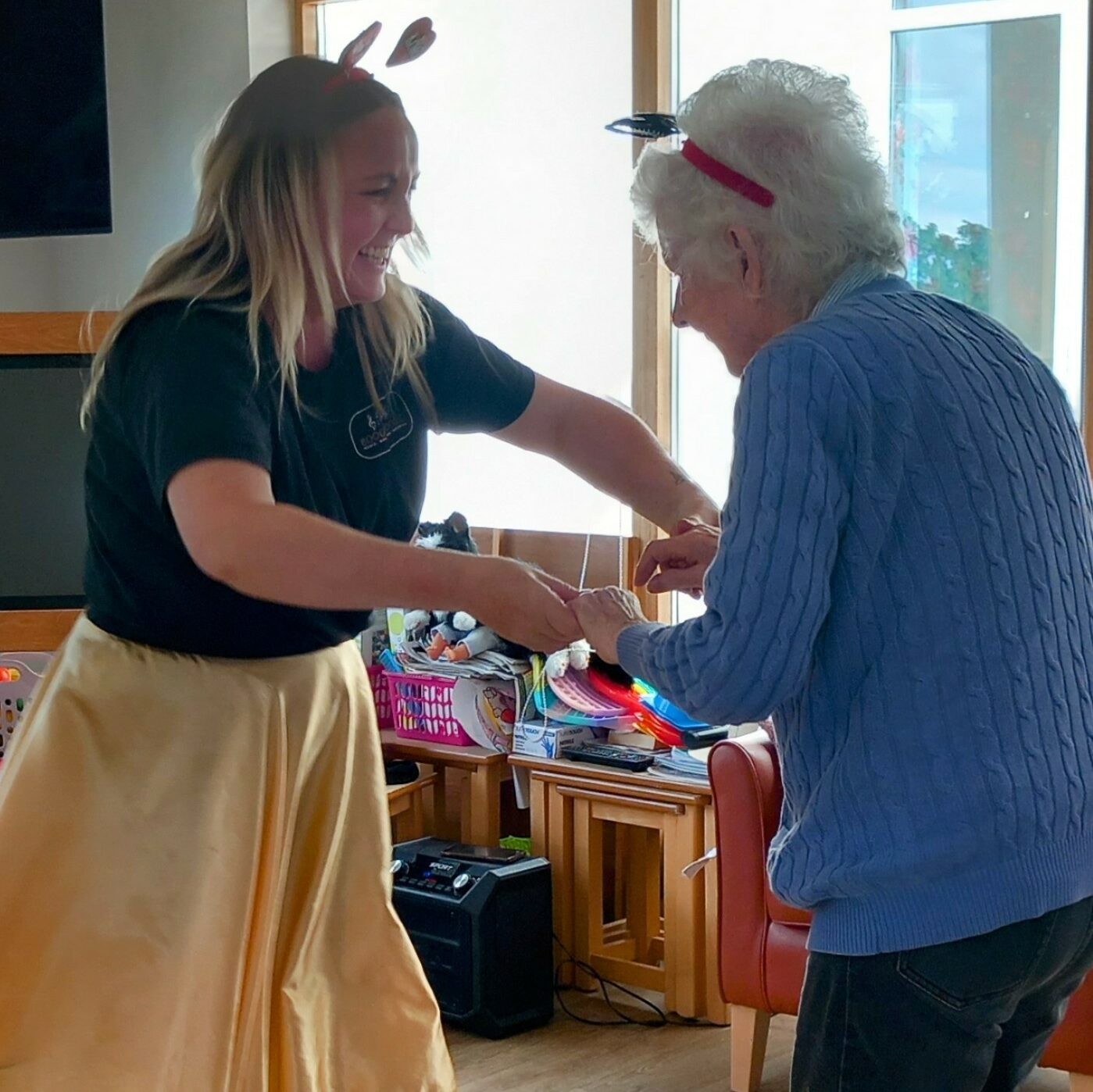 A younger woman and an older woman wearing headbands share laughter as they dance together in a room with wooden flooring and shelves of toys in the background.
