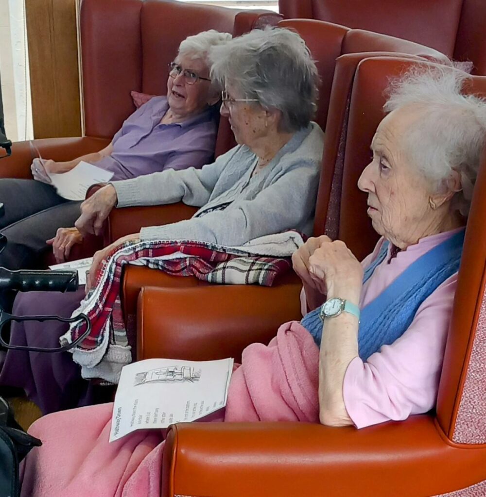 Three elderly women sit in red armchairs, blankets over their laps and papers in hand, as though taking a gentle stroll down Memory Lane in a warm communal indoor setting.