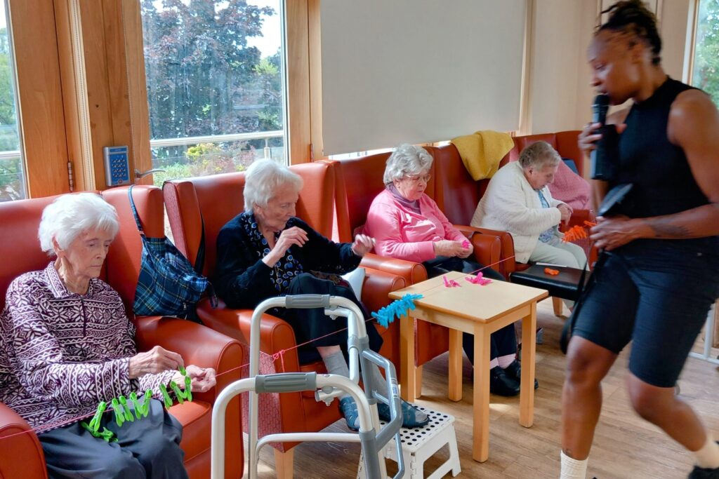 Four elderly people sit on red chairs in a common room, working with coloured pegs, while a woman stands nearby and speaks or sings into a microphone.