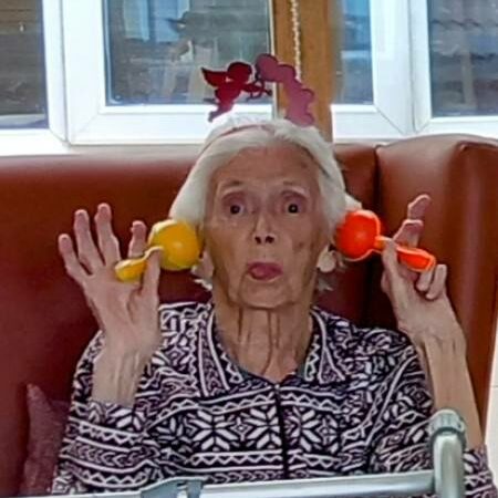 Elderly woman seated in a chair, holding colourful maracas to her head and pulling a playful face, sharing laughs as she wears a patterned jumper and a paper crown.