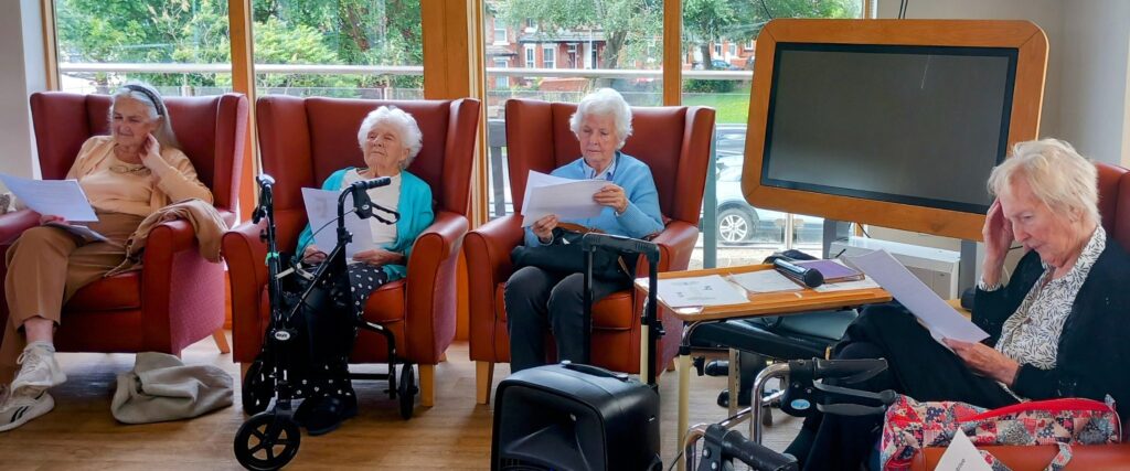 Four elderly women sit in armchairs, reading from papers; mobility aids are close by, and a large television screen is in the background, as though taking a quiet stroll down memory lane together.