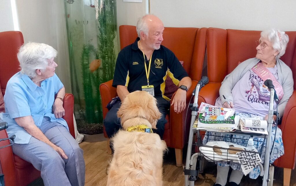 An older man and two elderly women sit and chat in a care home, with a Golden Retriever therapy dog in front of them and a walking frame holding magazines nearby.
