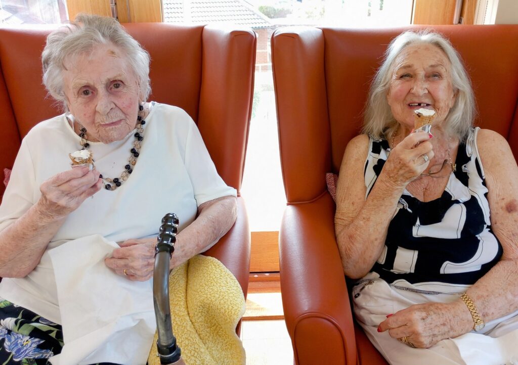 Two elderly women enjoy a pampering session, sitting in armchairs by a window, eating ice cream cones and smiling.