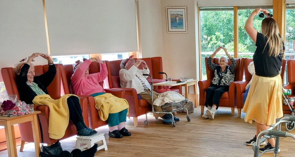 A group of elderly people seated in chairs follow a woman at the front, raising their arms overhead as they boogie together and share laughter in a bright, cheerful room.