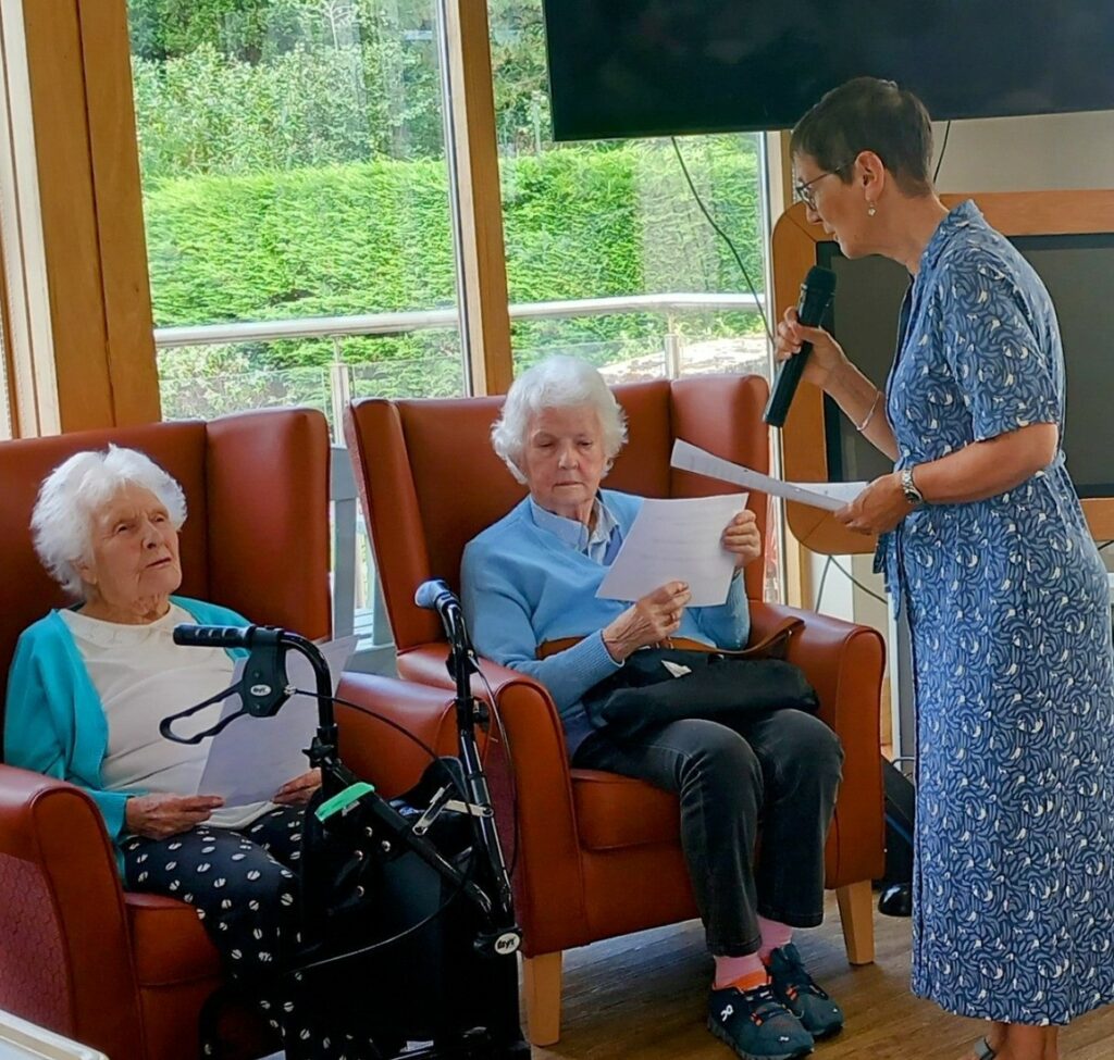 Three elderly women, two seated with papers and one standing with a microphone, take part in a Memory Lane activity indoors near large windows overlooking a tranquil garden.