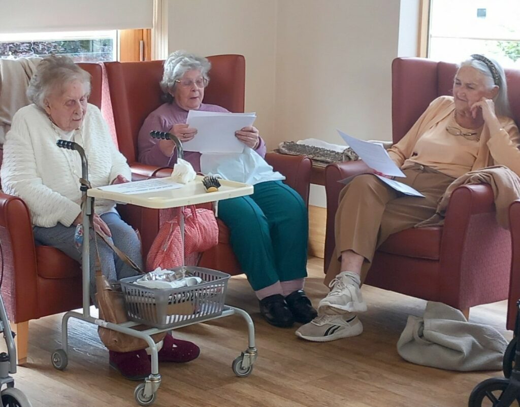 Three elderly women sit in armchairs indoors, reading from sheets of paper. A walking frame and a tray table with various items are nearby. The tranquil scene exudes nostalgia as they take a gentle stroll down memory lane together.