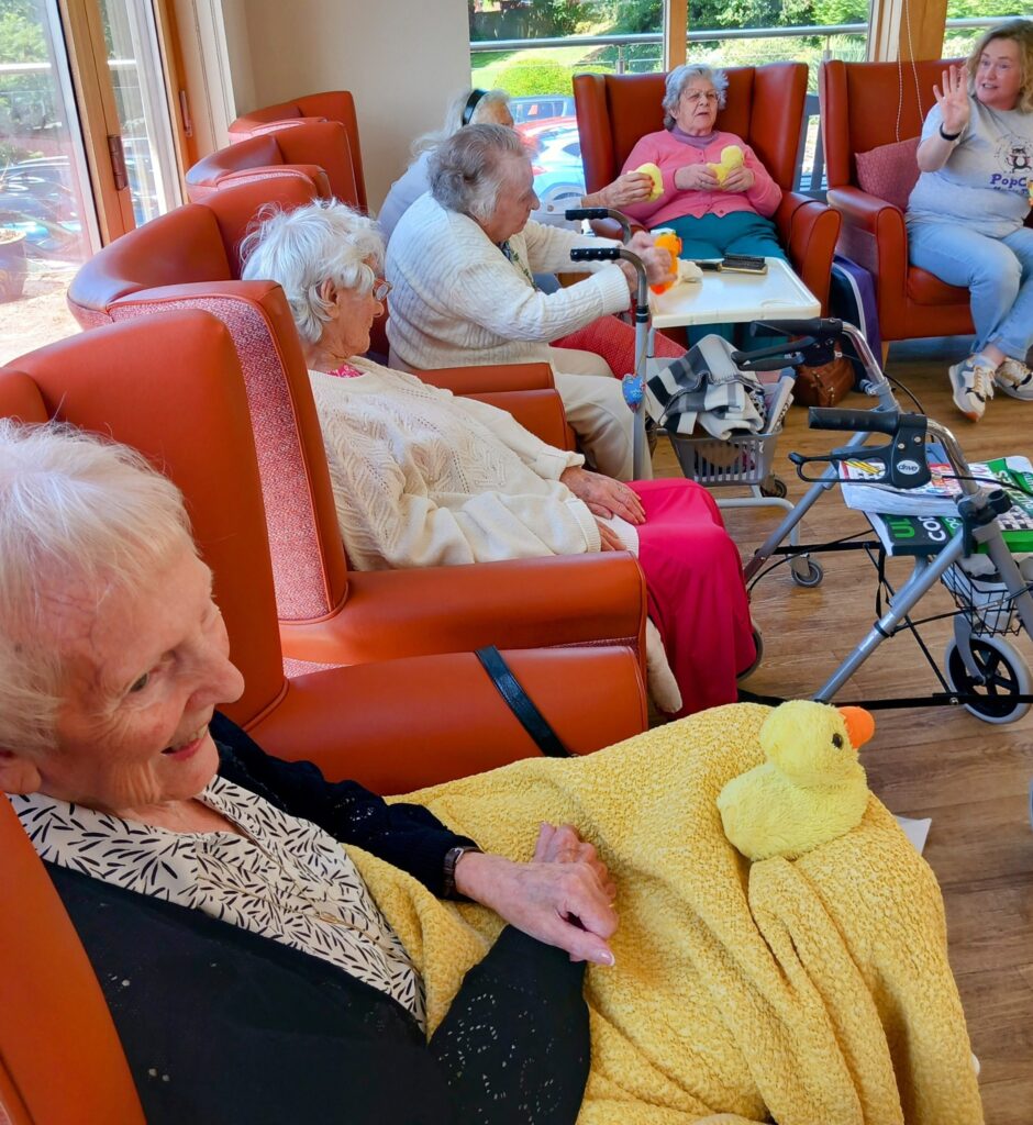 A group of elderly women sit in orange chairs in a bright room, some holding soft toy ducks, as a younger woman engages with them, bringing generations together.