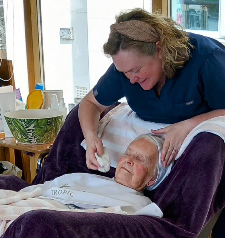 A woman in a navy shirt gently wipes the face of an elderly woman reclining in a chair covered with a towel, enjoying a soothing pampering session in a well-lit room.