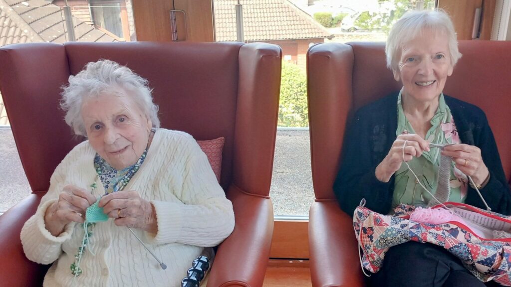 Two elderly women sit in armchairs by a window at Abbeyfield Lear House, knitting with pastel-coloured wool and smiling at the camera.