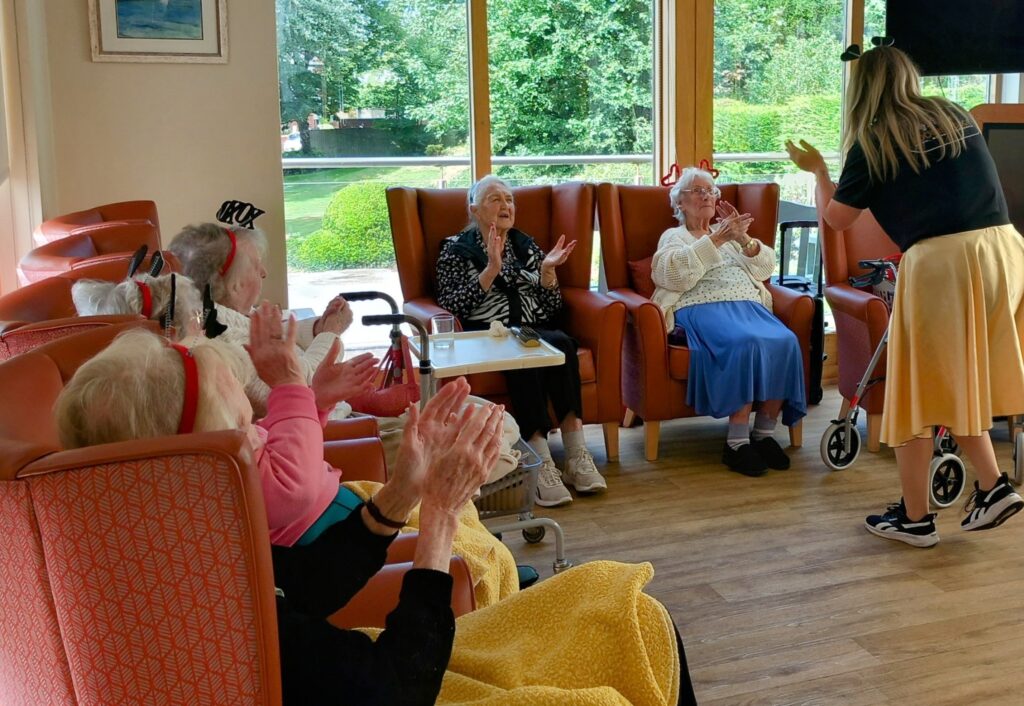 A group of elderly people sit in armchairs, clapping and smiling affectionately, while a woman stands before them leading a joyful activity in a bright, spacious room filled with laughter and large windows.