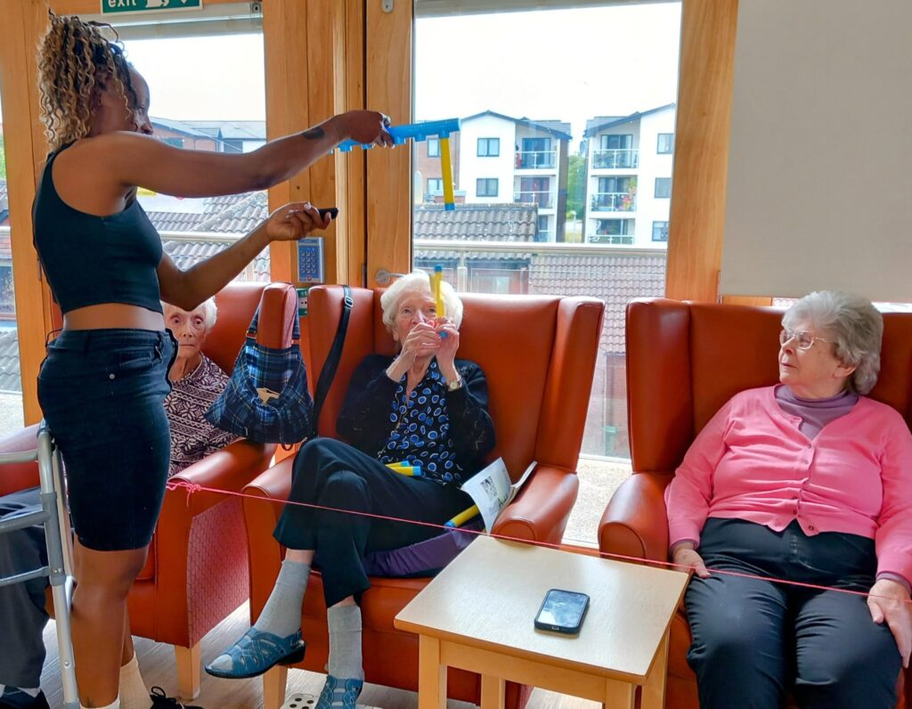 A woman holds a toy fishing rod towards three elderly women seated in armchairs in a brightly lit room, with a phone and string visible in the foreground.
