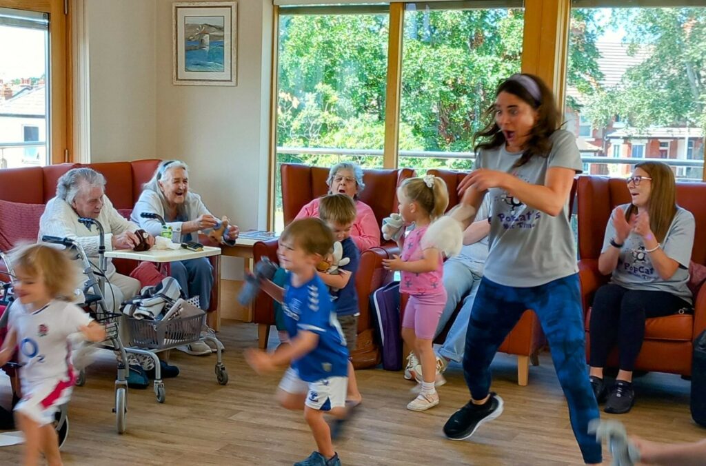 Children and adults interact energetically in a brightly lit room, bringing generations together as some pensioners sit on a sofa while others join in the activities.