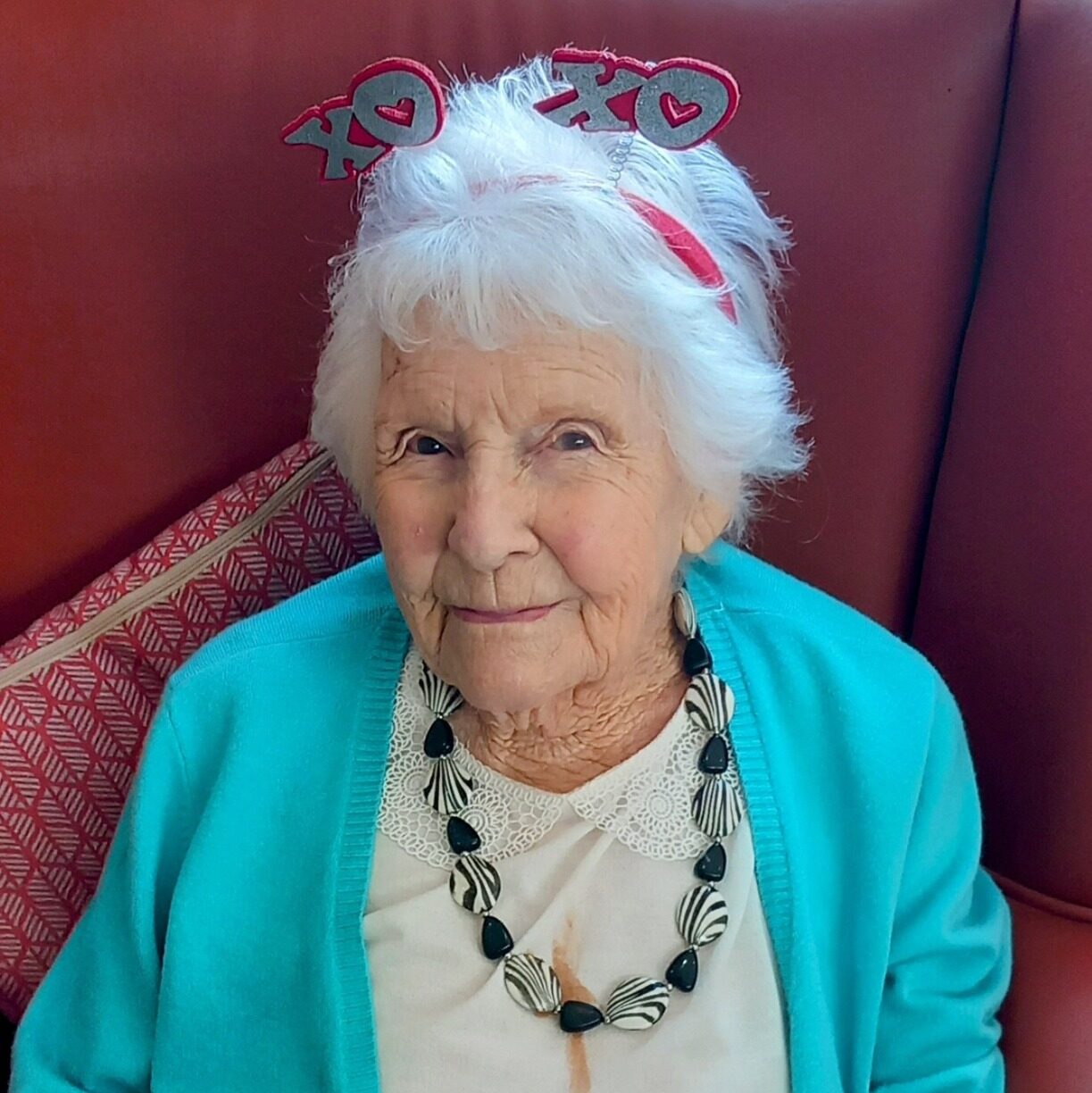 Elderly woman with white hair, wearing a turquoise cardigan, a shell necklace, polka-dot trousers, and a red “XO” headband, seated in a red chair with her hands folded, radiating love and gentle laughter.