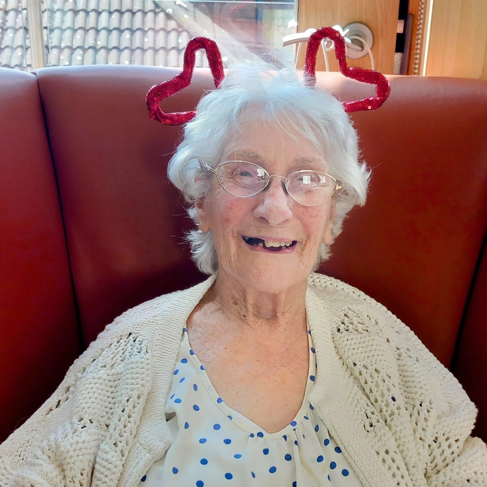Elderly woman with glasses sits on a red chair, wearing a white polka-dot top, cream cardigan, and red heart-shaped headband, spreading love and laughter with her charming smile.