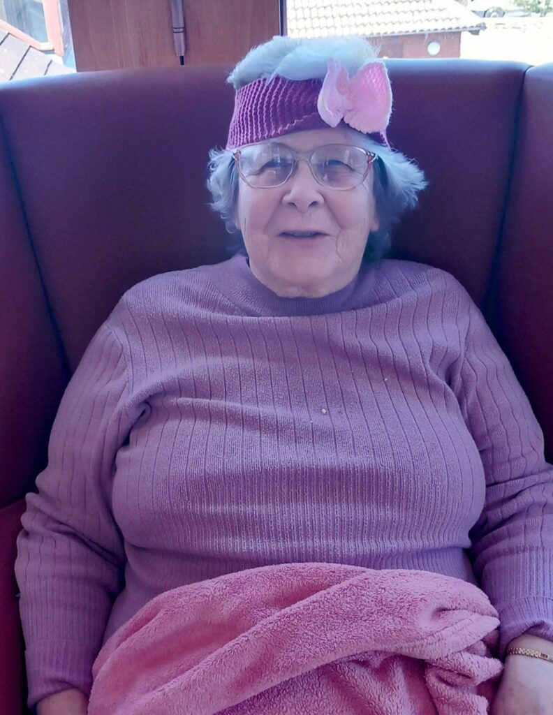An elderly woman with glasses is seated on a chair at Abbeyfield Lear House, wearing a purple jumper and a matching headband adorned with a pink bow. She is knitting, while a pink blanket rests on her lap.